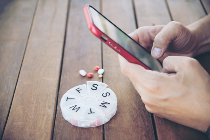 Hände tippen auf Smartphone neben Tablettenbox mit Pillen