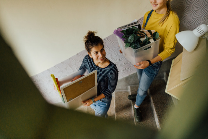 Zwei junge Frauen tragen Umzugskarton eine Treppe hinunter.