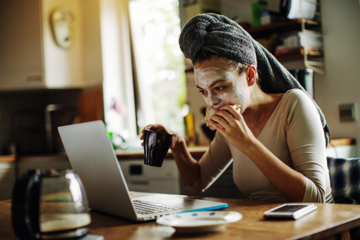 Frau mit einer Pflegemaske im Gesicht isst und arbeitet gleichzeitig am Laptop