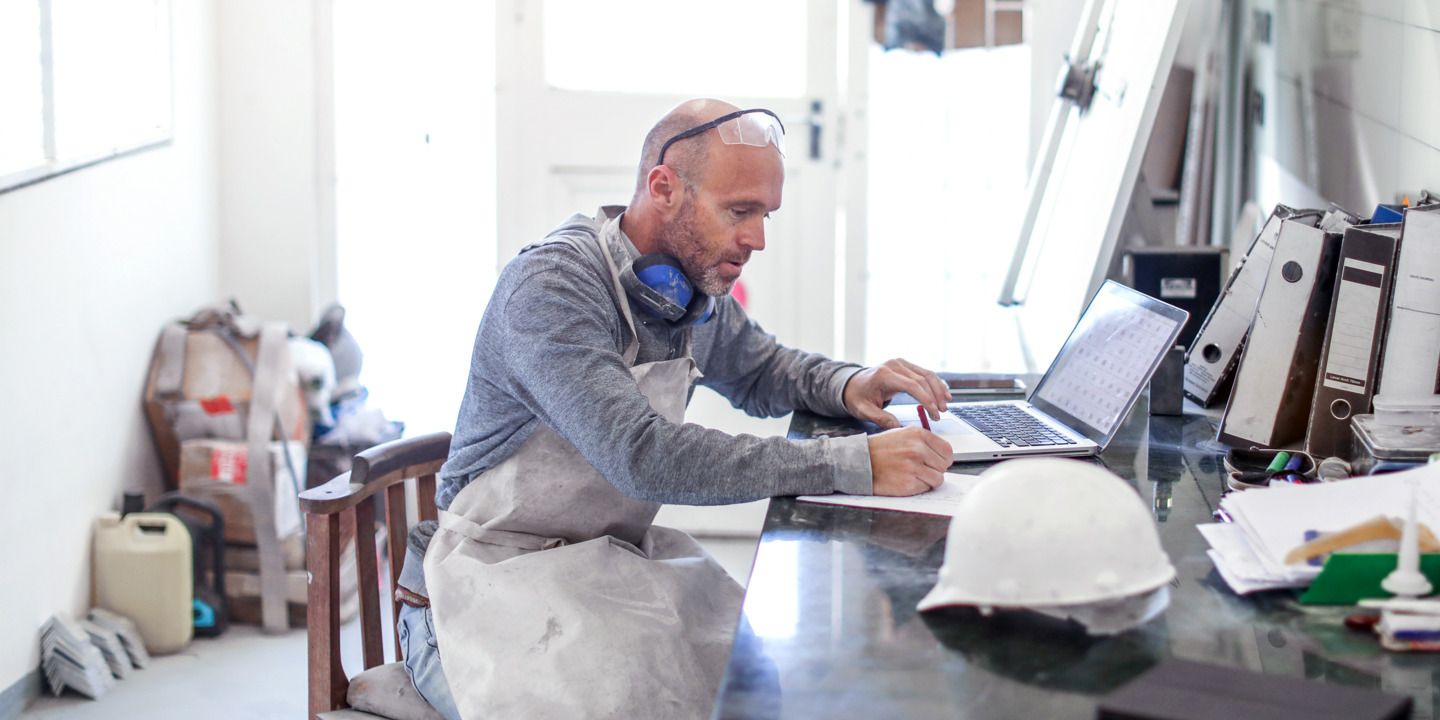 Ein Mann in Arbeitskleidung sitzt in seiner Werkstatt und benutzend einen Laptop schreibt er etwas auf einem Papierblatt.