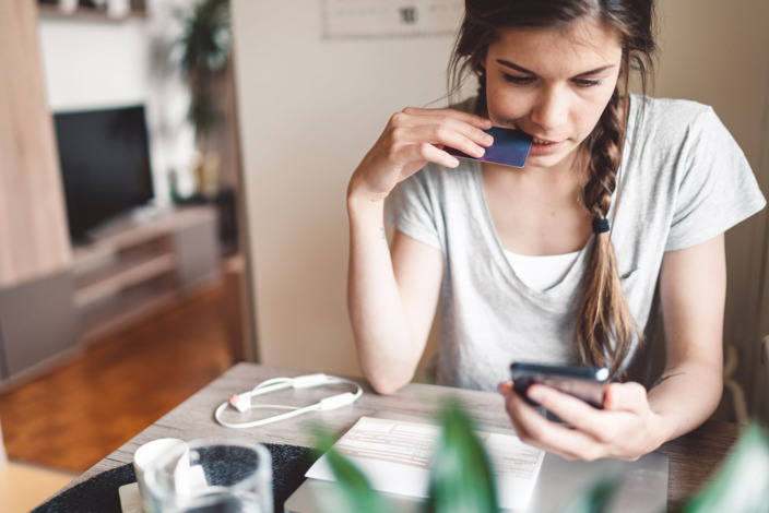 Junge Frau beißt leicht in ihre Kreditkarte und schaut unglücklich auf ihr Handy.