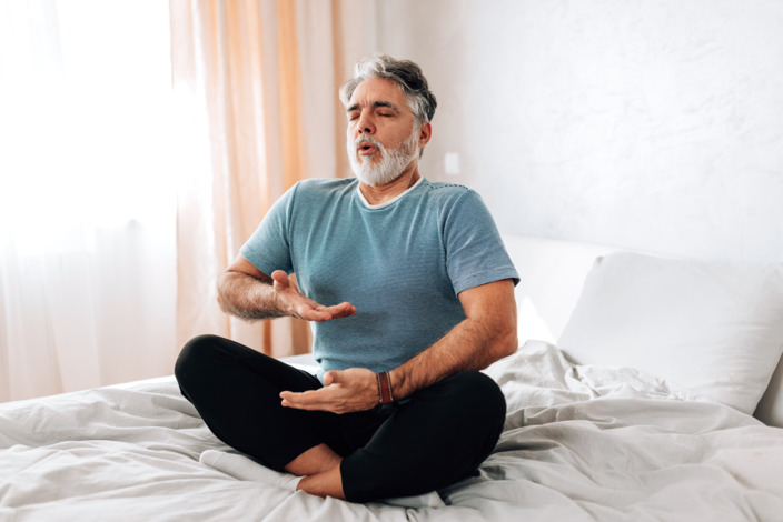 Ein Mann sitzt auf dem Bett und macht Atemübungen. 