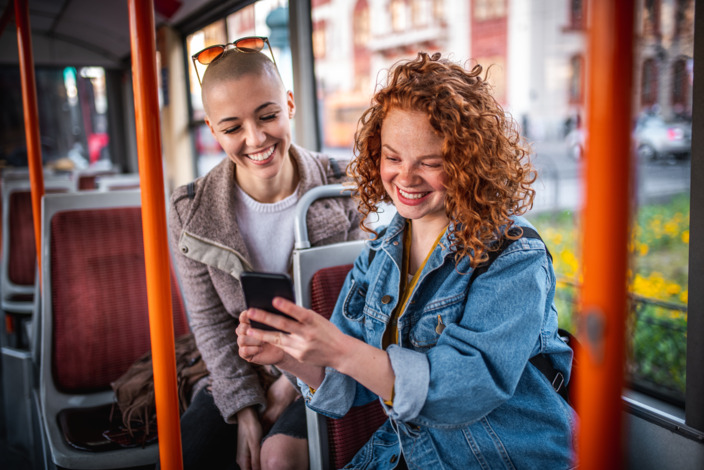 Zwei junge Frauen sitzen im Bus und schauen lachend auf ein Smartphone.