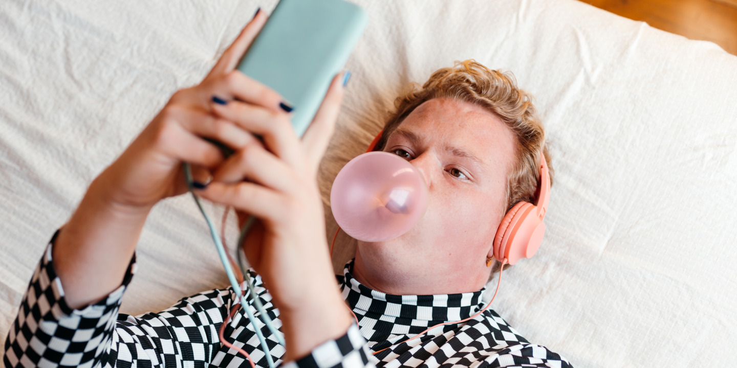 Junger Mensch, der mit Smartphone in der Hand auf dem Bett liegt und eine Kaugummiblase macht.