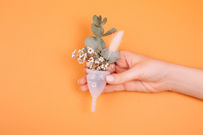 Weiße Menstruationstasse mit Blume in der Hand der Frau auf orangem Hintergrund