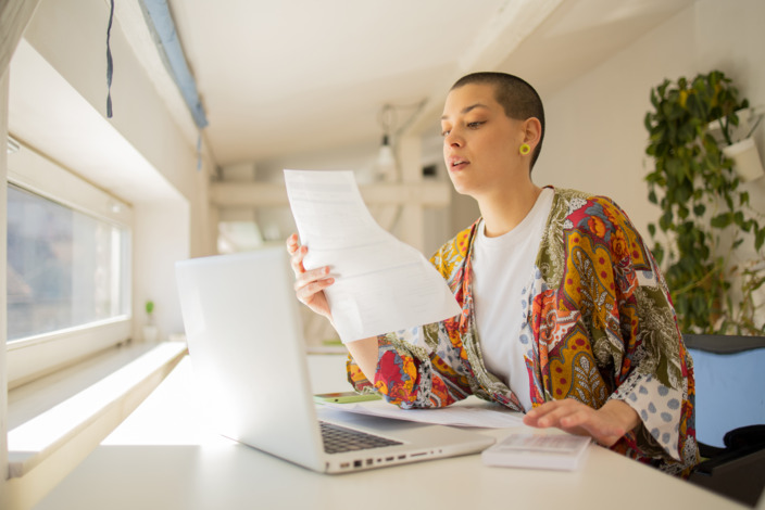 Junge Frau sitzt mit Laptop am Schreibtisch und liest ein Schreiben in der Hand