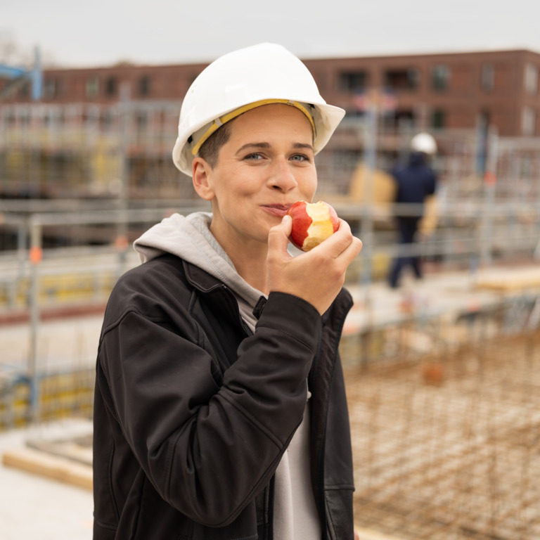 Frau mit Helm auf Baustelle isst Apfel