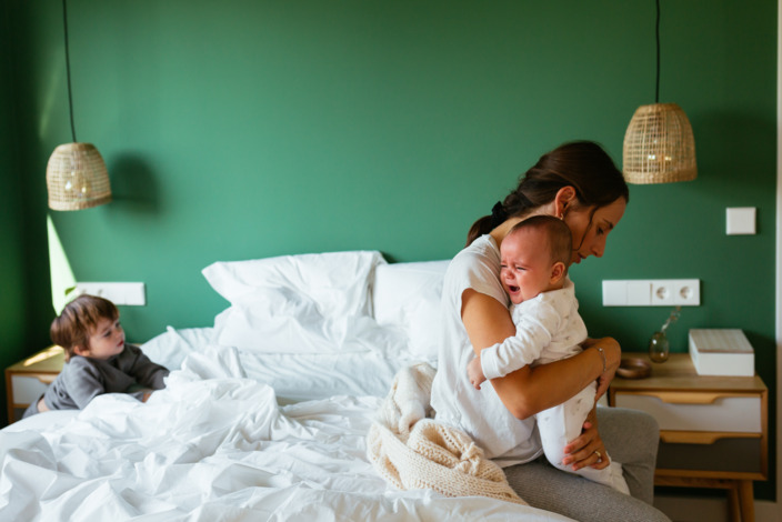 Mutter sitzt erschöpft auf dem Bett und tröstet ihr weinendes Baby, der zweijährige Bruder schaut zu