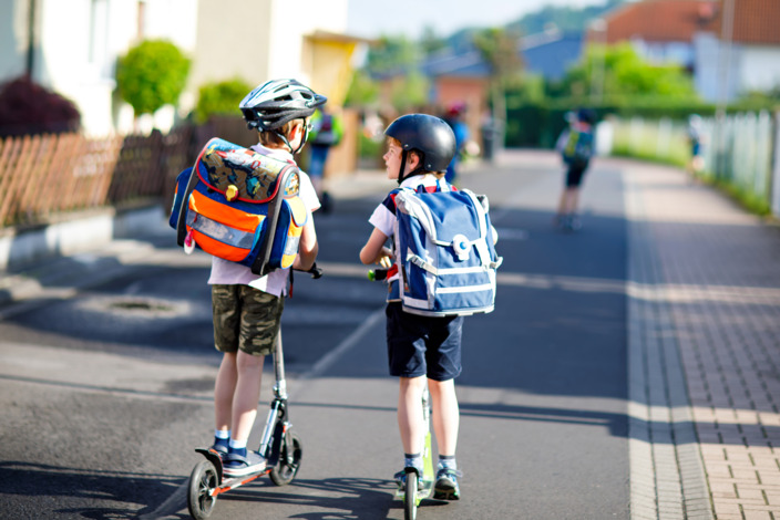 Zwei Grundschüler mit Schulranzen auf dem Rücken fahren mit dem Tretroller zur Schule