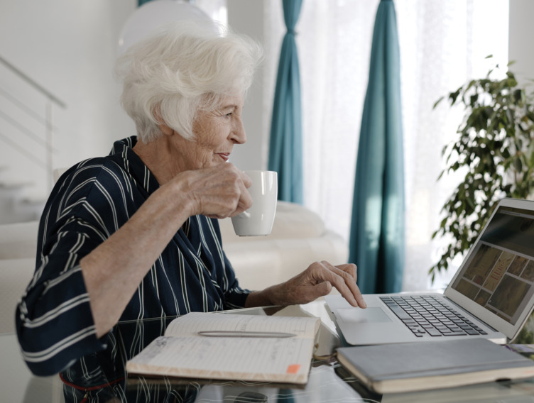 Seniorin sitzt mit einer Tasse Tee vor dem Laptop und prüft ihre Unterlagen