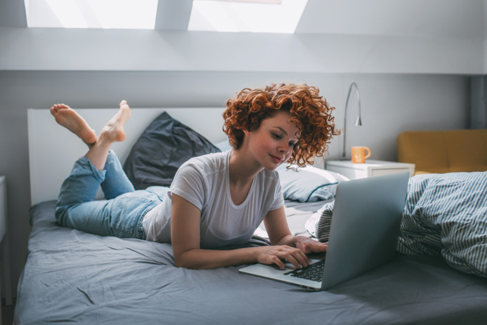 junge Frau liegt auf ihrem Bett und benutzt ihren Laptop